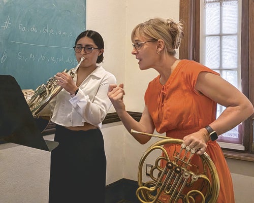 Photo of horn professor holding a french horn while instructing a student that is playing a french horn