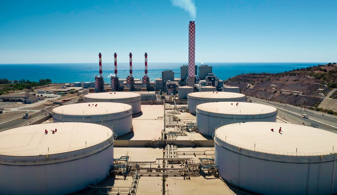 Aerial view of power station featuring large white fuel storage tanks and red chimneys emitting smoke along the Cyprus coastline near the Mediterranean Sea.