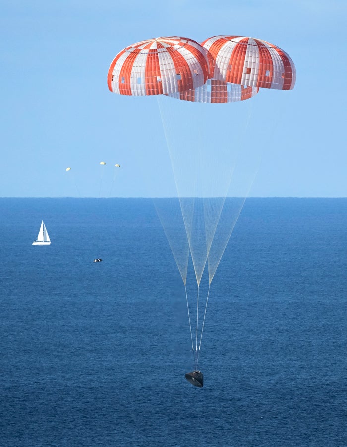 Photo of the Orion capsule landing over the sea with three red and white striped parachutes above. The ocean includes a sail boat further off in the distance.