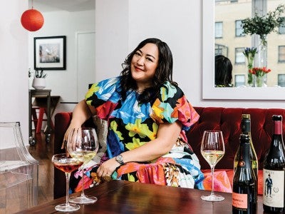 Photo of a smiling Belinda Chang sitting on a red velvet sofa with wine bottles on the table in front of her