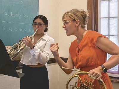 Photo of horn professor holding a french horn while instructing a student that is playing a french horn