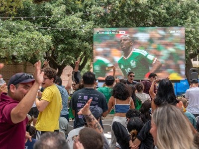 Photo of watch party outside in the courtyard of Valhalla with fans cheering for Mexico in front of a large tv screen showing the game