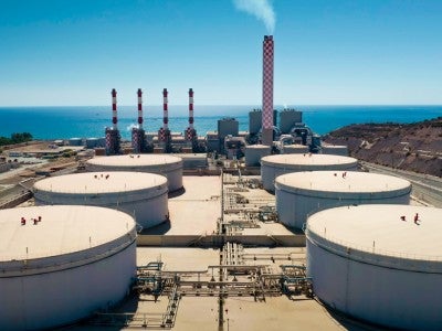 Aerial view of power station featuring large white fuel storage tanks and red chimneys emitting smoke along the Cyprus coastline near the Mediterranean Sea. 
